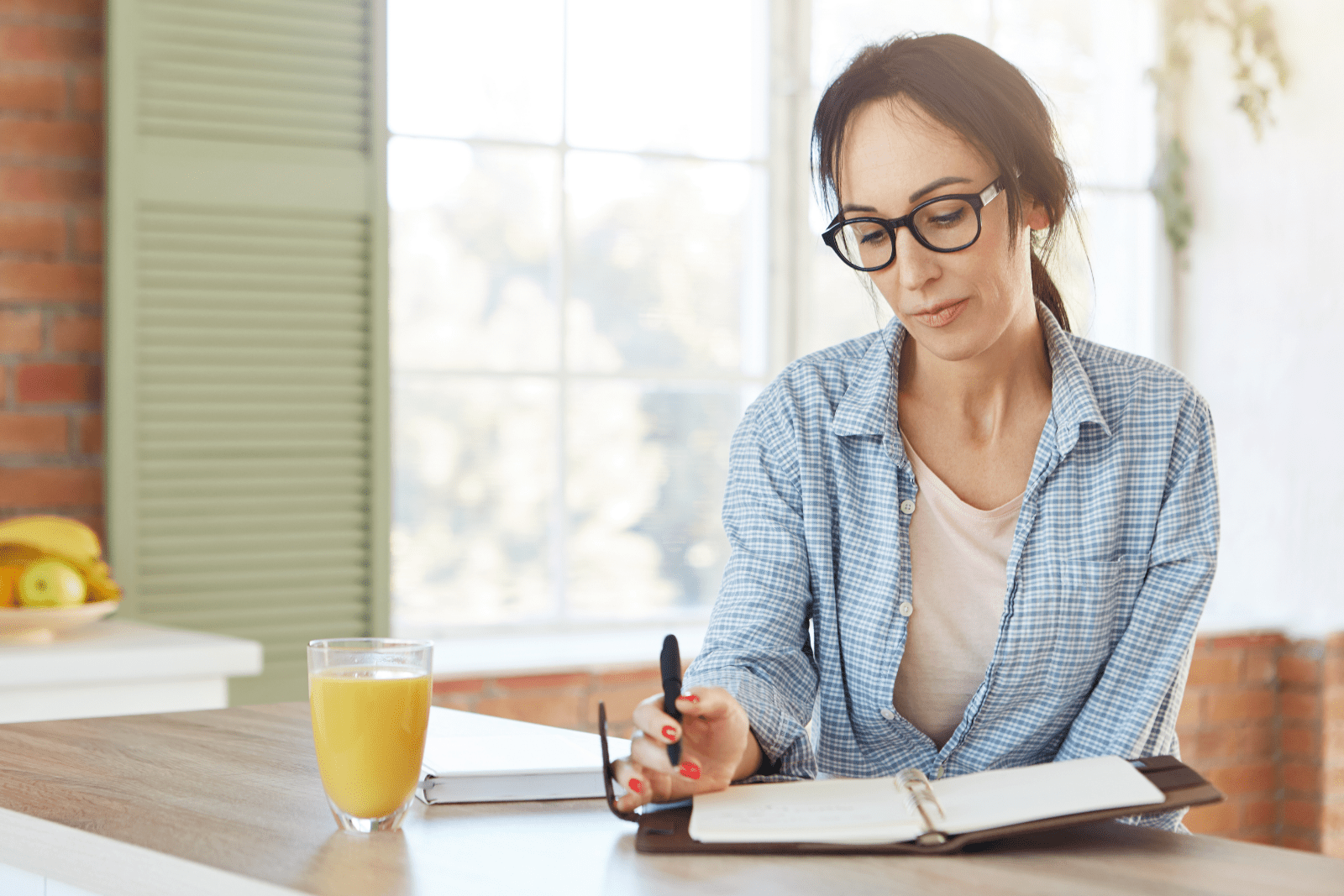 Woman sitting at a kitchen table with an open planner, reflecting on her daily routine in natural morning light.