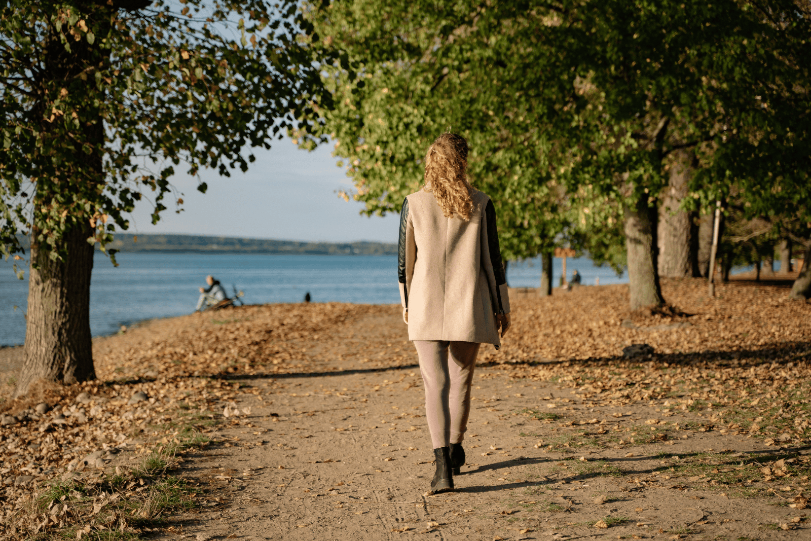 Back view of a woman walking along a quiet lakeside path surrounded by autumn trees.