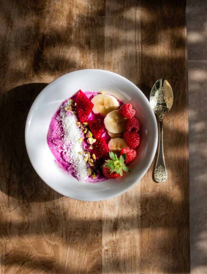 Overhead close shot of a dragon fruit frozen yogurt bowl with banana, raspberries, strawberry, toasted coconut and pistachio, served with a vintage spoon on a wooden table
