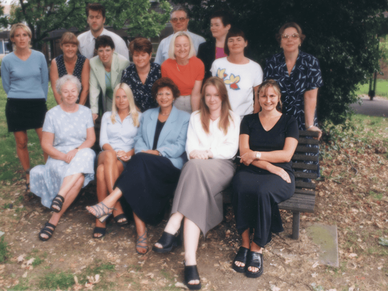 A group of people posing for a photograph around a park bench. 5 people are sat on the bench, with another 11 people stood behind it.