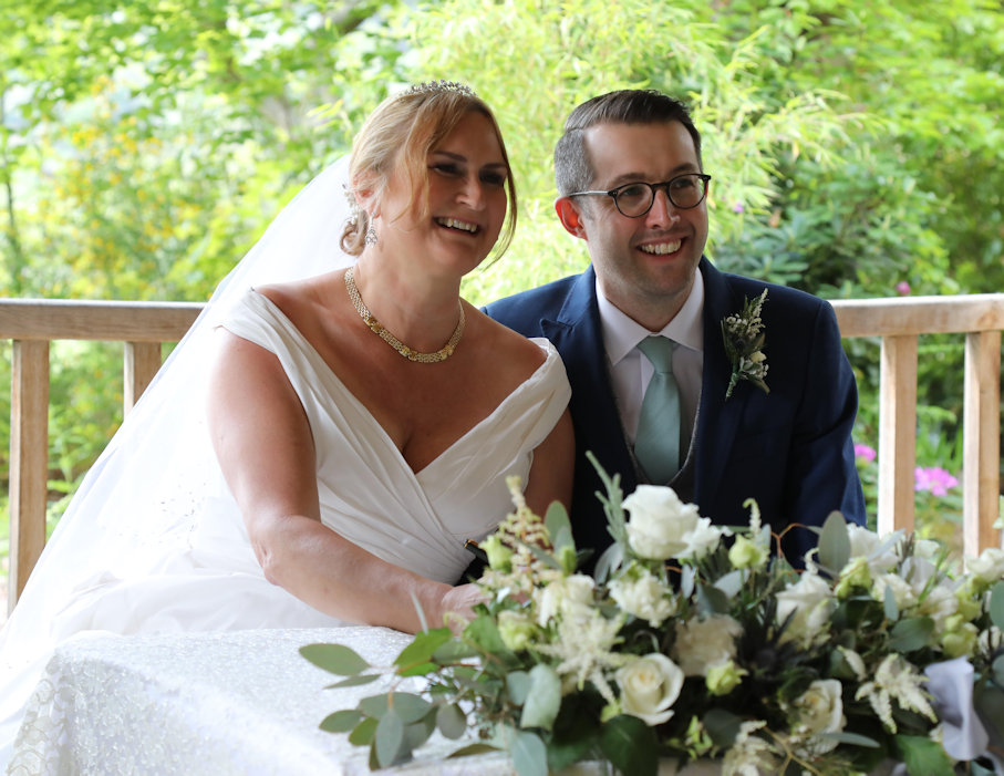 A man and a woman on their wedding day, sat at a table, smiling and signing a book in front of them with a bouquet of flowers.
