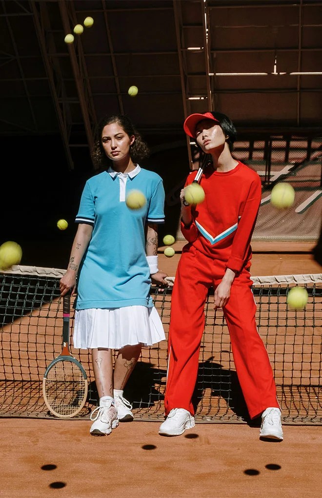 Photograph of two models posing in a Tennis court. On the left, the model is wearing a blue polo shirt with a white skirt holding a tennis racket, on the right the model is wearing a red jumper. Tennis balls are flying.