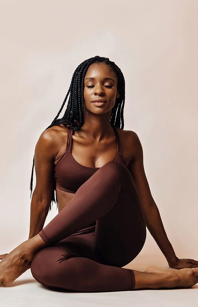 Photo of black model wearing a brown, yoga suit stting down on a light brown backgorund.