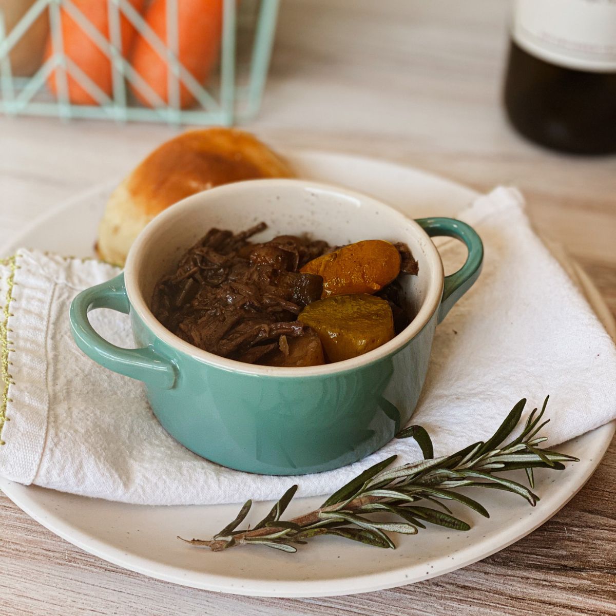 Bowl of pot roast on a plate next to a sprig of rosemary and a dinner roll. Pot roast is on front of a basket of carrots and a bottle of wine.
