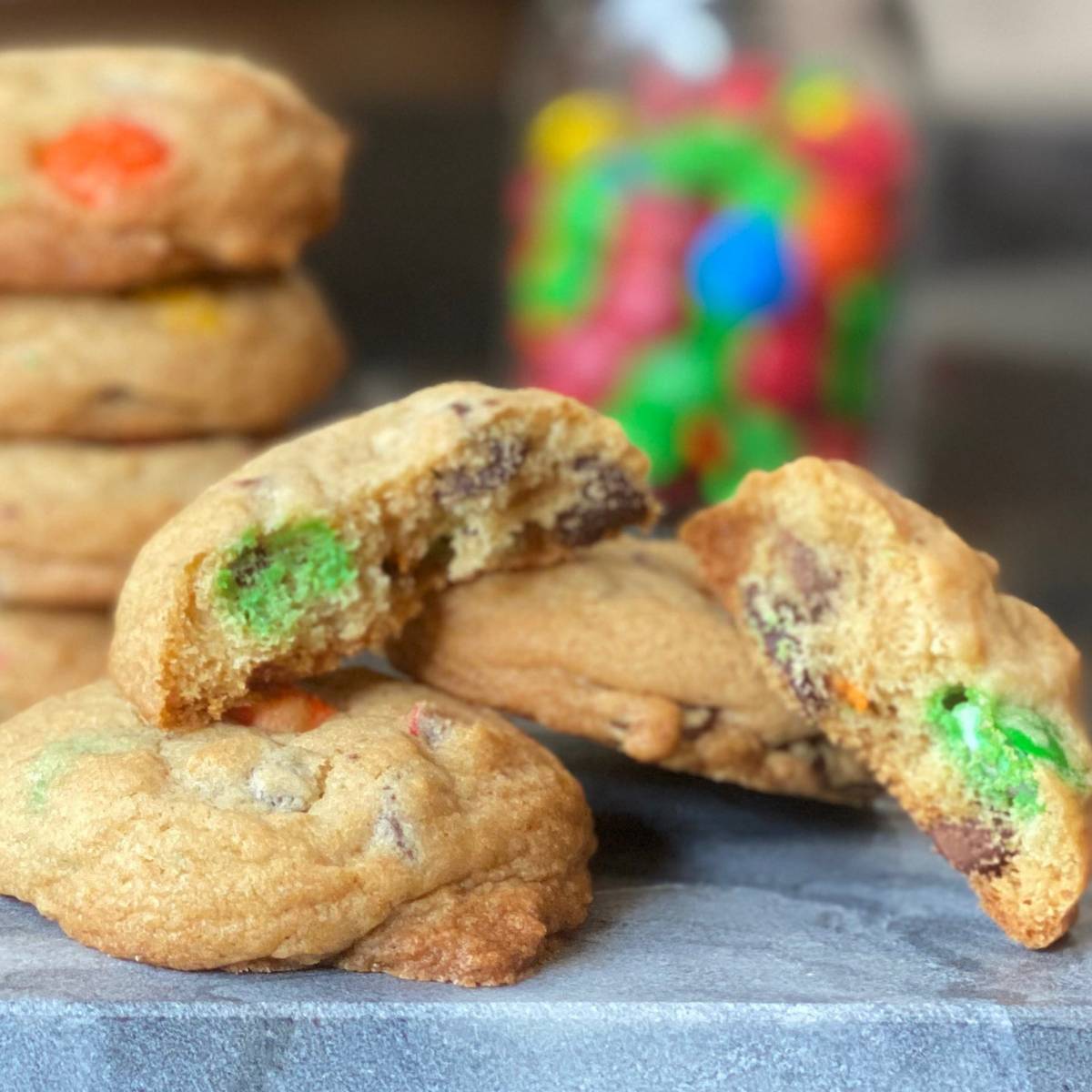 M&M cookie broke in half sitting on top of two whole cookies in front of a stack of cookies and a jar of M&M candy.