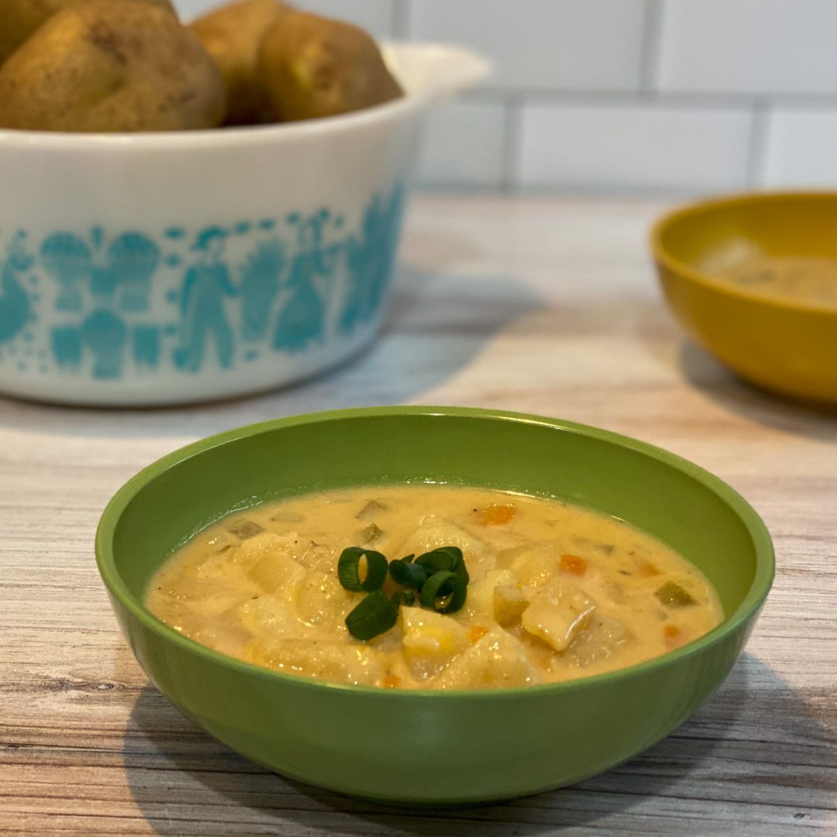 Bowl of potato soup garnished with green onions in front of a bowl of raw potatoes and another bowl of soup.