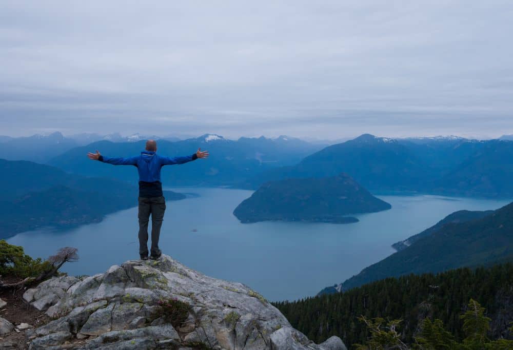 Man standing on mountain arms spread wide in front of a mountain range that is divided by a large lake. Showcasing solo travel for introverts.