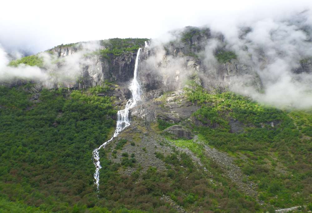 Long waterfall in Norway from a tree covered mountain side with clouds lingering before the wall.