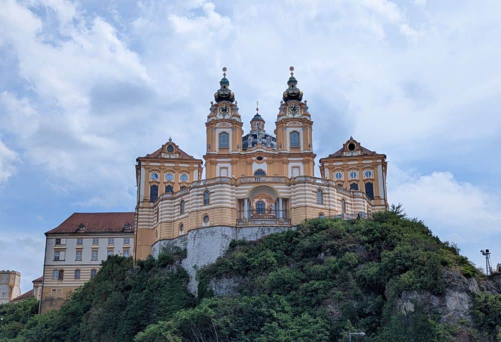 Melk Abbey from the lower level of the town. Golden coloured building with two spires. Green trees in front of the mountain on the lower half of the picture