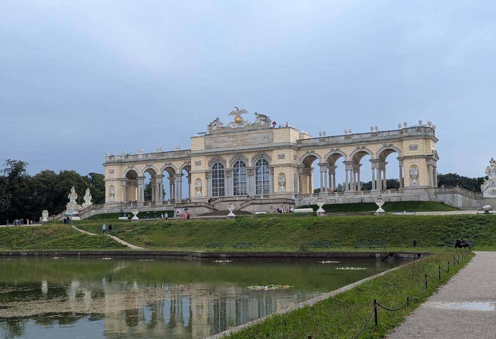 View of Gloriette with lake in front and grass around it. Slightly grey sky in the back.