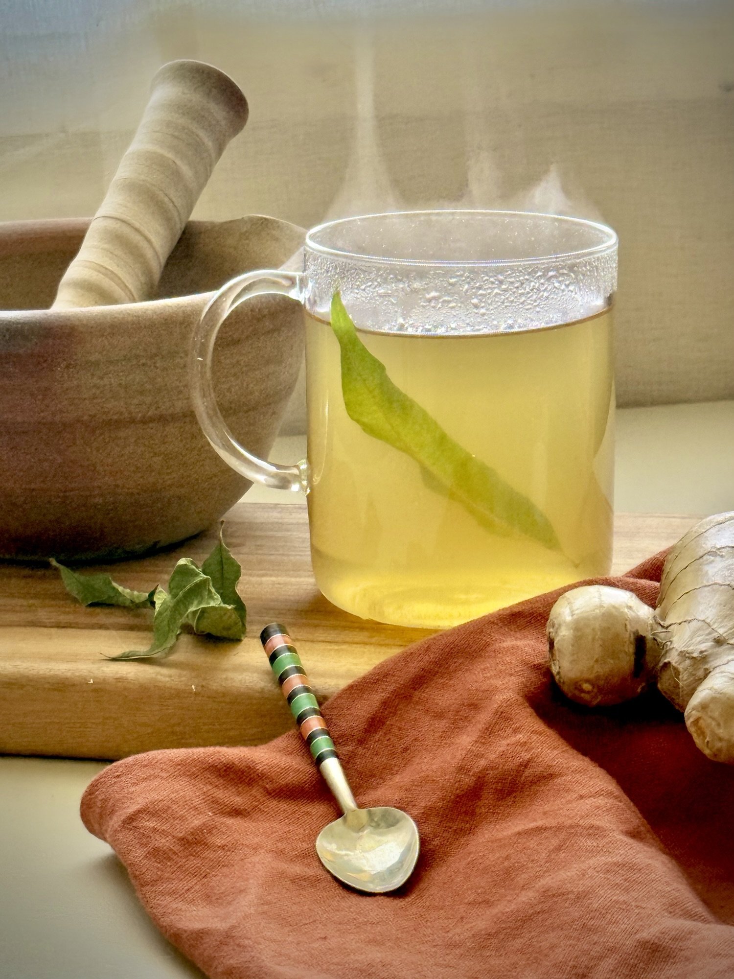 A steaming glass mug of pale yellow digestive seed tea,rests on a wooden board, a ceramic mortar and pestle is in the background, a striped spoon, ginger root and rust coloured tea towel in the foregorund. A single green leaf of lemon verbena is suspended in the tea.