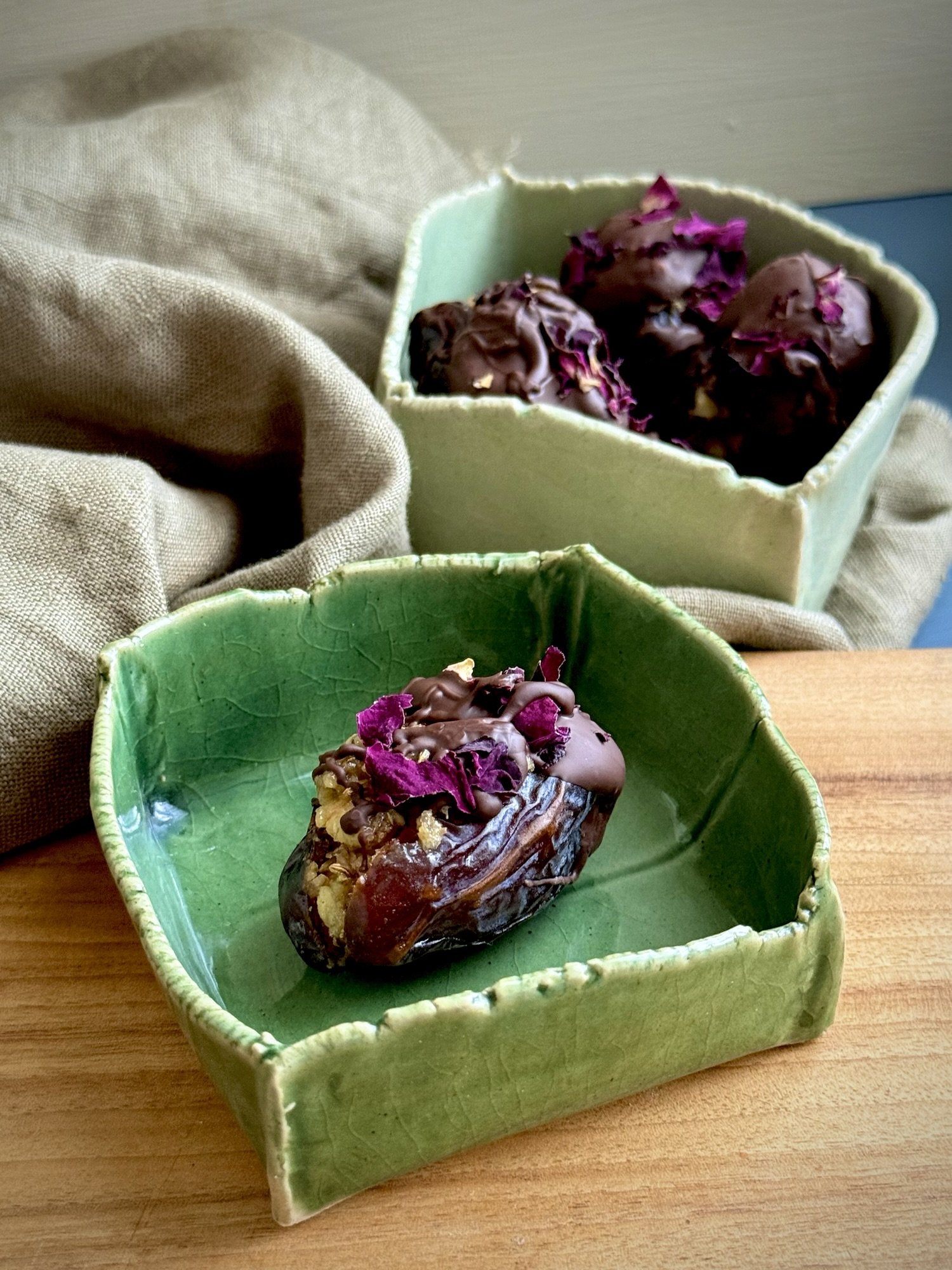 After Dinner Digestive Support Dates - Chocolate-drizzled stuffed Medjool dates topped with dried rose petals, served in handmade green ceramic dishes on a wooden table.