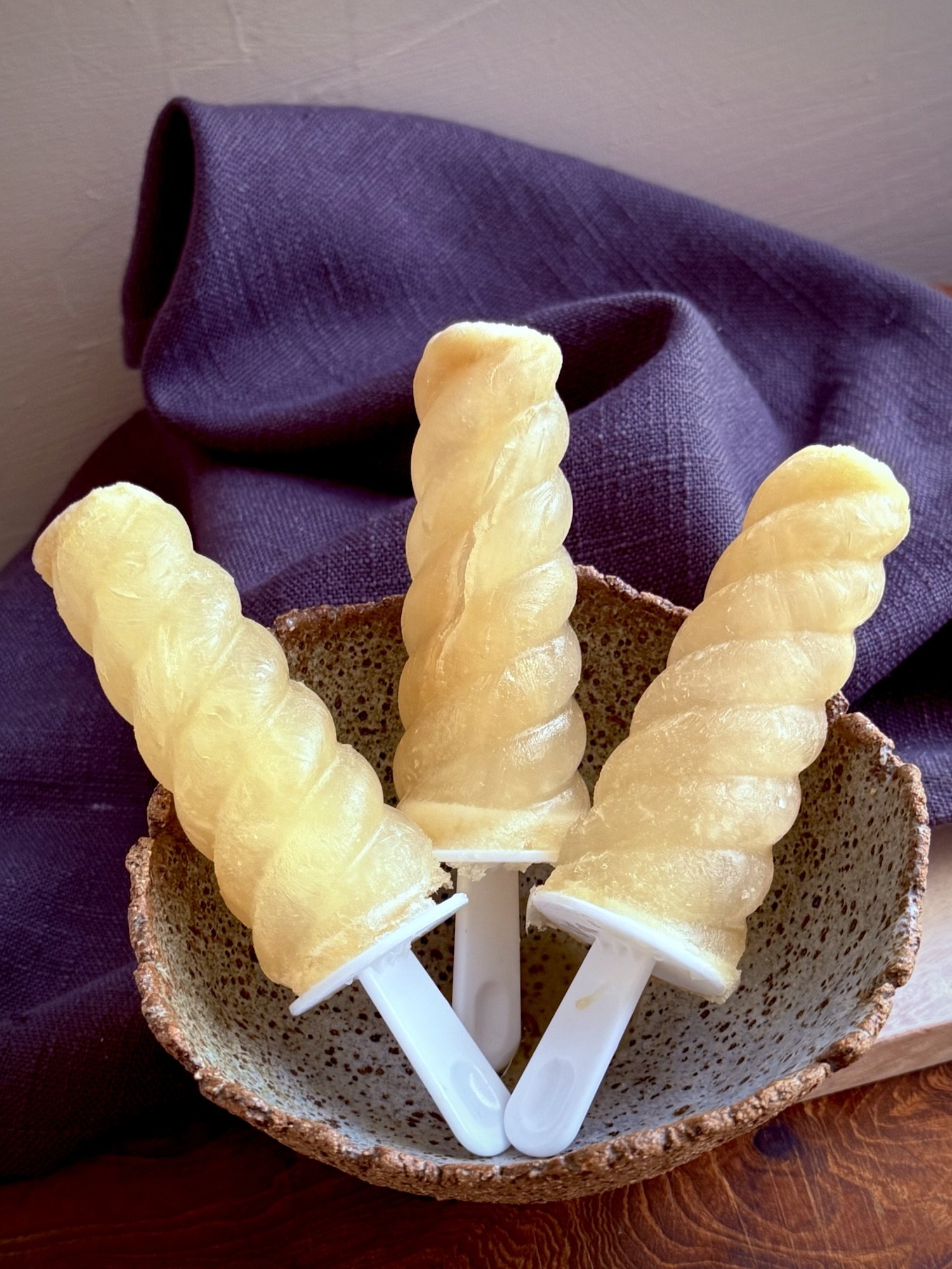 Three pale ginger and coconut-water anti-nausea icy poles in white moulds, standing in a rustic speckled ceramic bowl against a soft purple linen cloth.