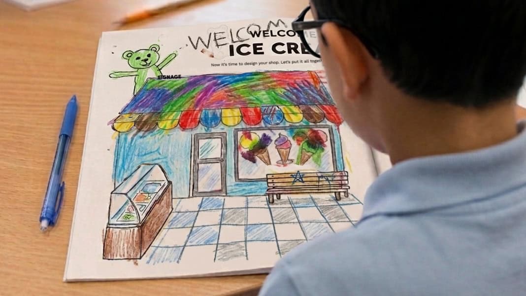 A Chinese student sitting at a desk, drawing with a pen inside his open "You Made It!" activity workbook by The Mind Orchard.