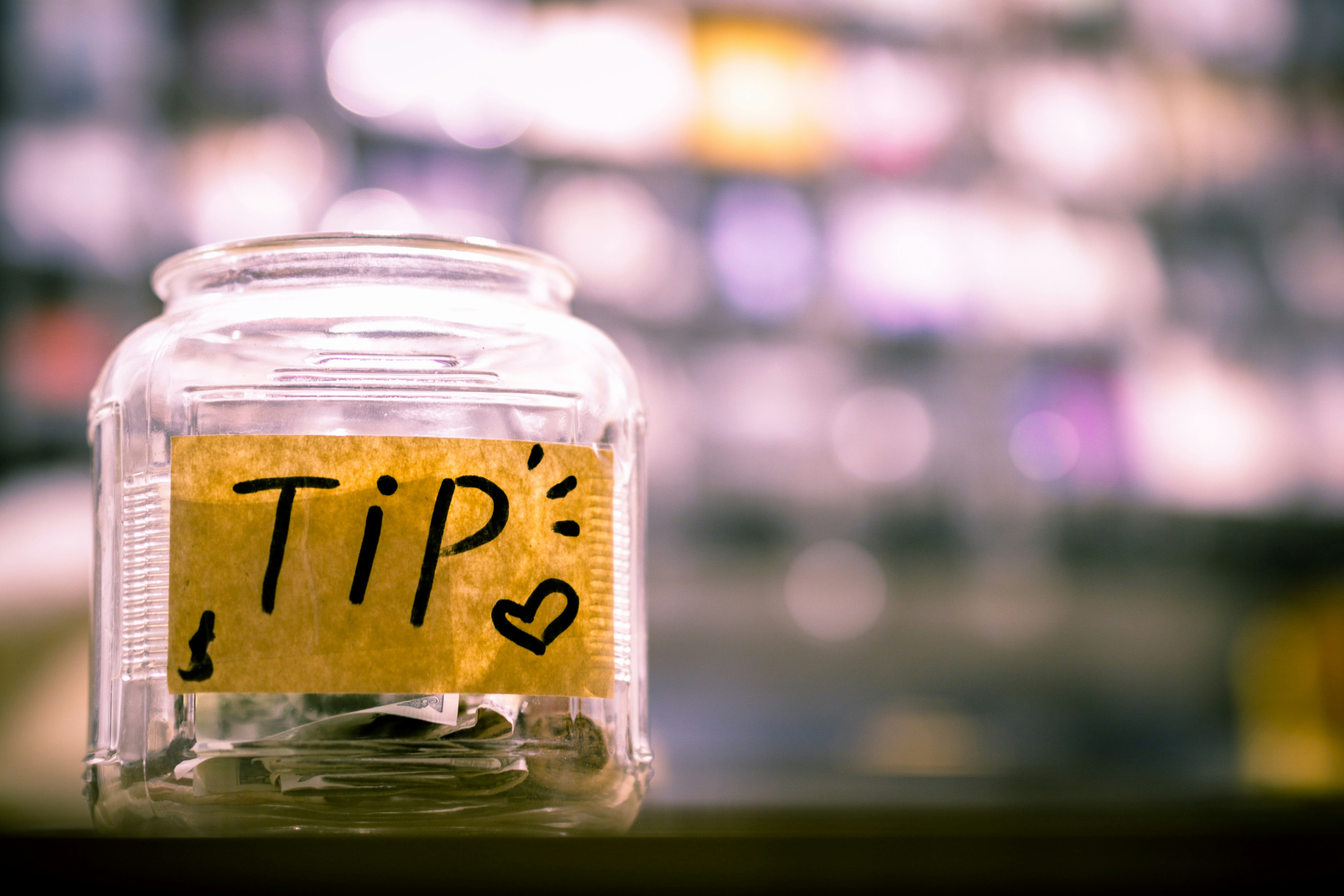 Glass tip jar with handwritten “TIP” sign and heart symbol, containing cash, against a blurred colorful background.