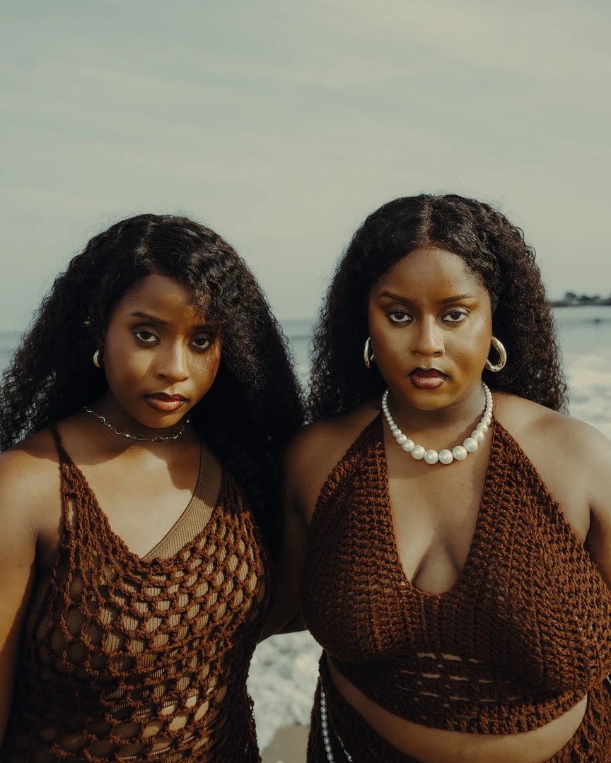Oiza and Meyi, Nigerian twin duo, standing together on a beach wearing matching brown crochet outfits with ocean backdrop