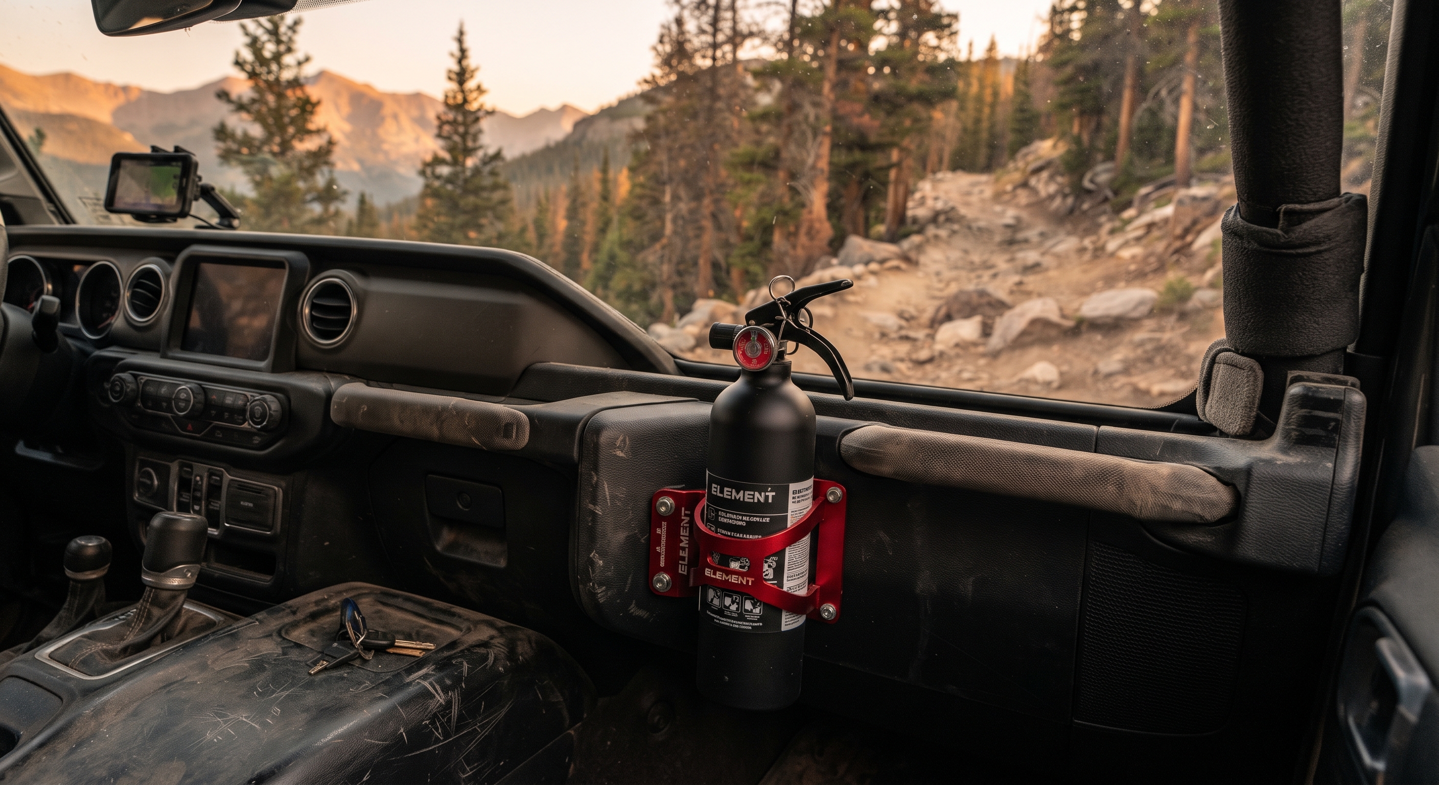 Interior view of a dusty Jeep Gladiator cabin with a matte-black Element fire extinguisher securely mounted on the passenger side of the center console; a winding mountain trail is visible in the blurred background through the windshield.