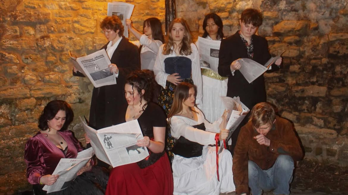 Nine actors, some standing and some sitting, are reading newspapers in a dimly lit room