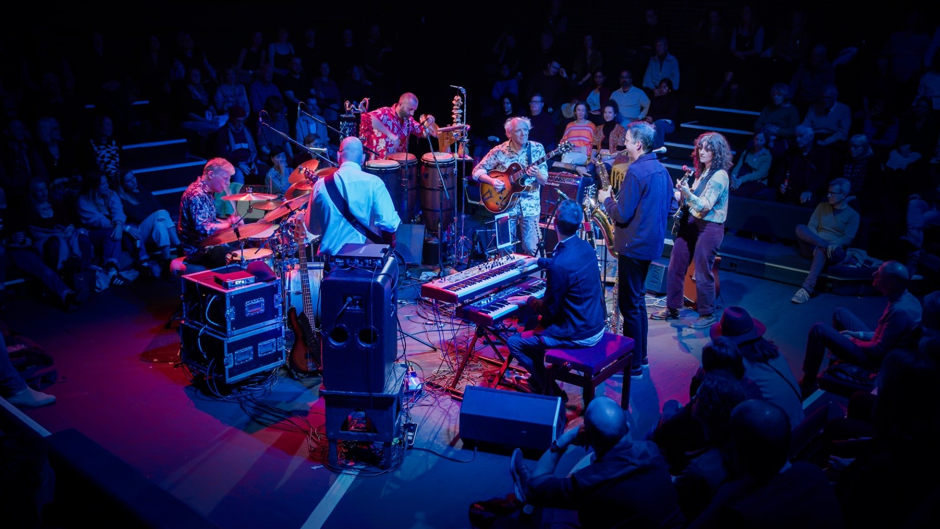 The seven members of Hejira perform, standingin a circle, on a stage lit in blue and pink hues.