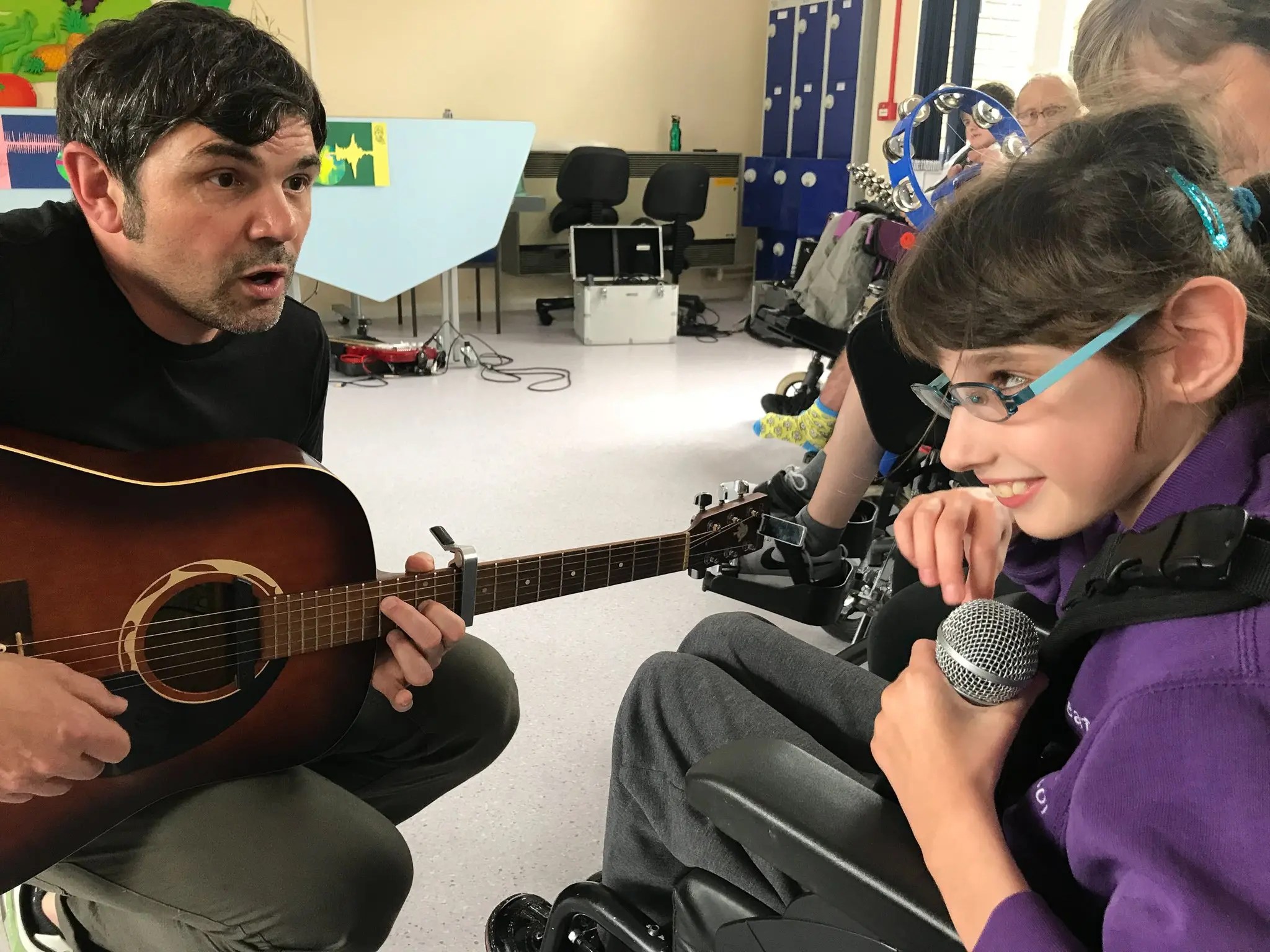 darts musician, Moony, sits with a guitar sits cross legged on the floor next to a girl with long hair and glasses singing into a microphone - a darts in Doncaster project
