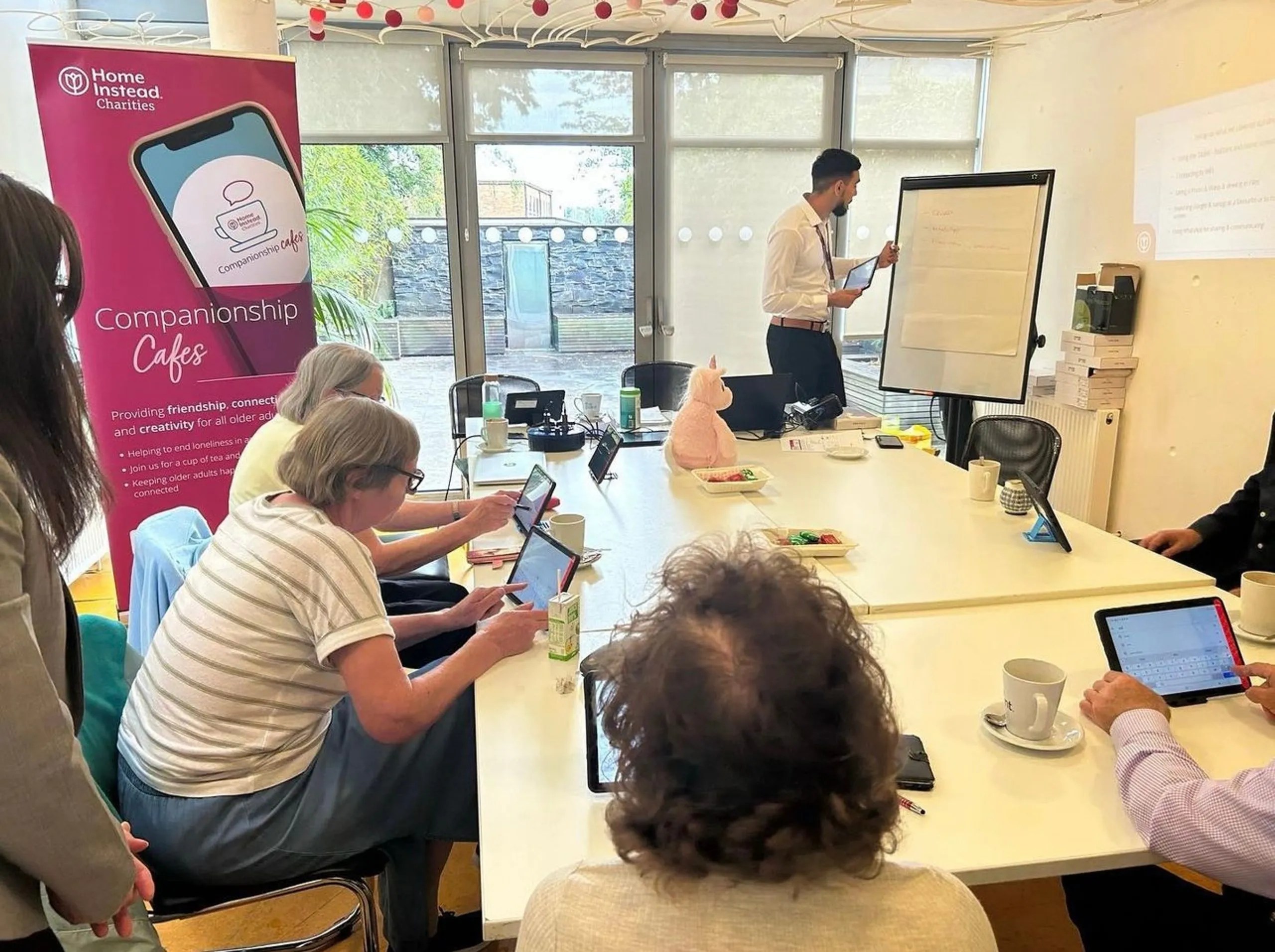 A group of older adults are seated around a large white table in the centre of a small, bright room. There are refreshments on the table and the adults are using tablets. Another adult is stood to the right of the table and is writing on a flip chart. A large pink banner on the left is advertising Companionship Cafes.
