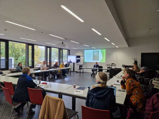 A photograph of the entire conference room, showing the participants engaged in discussion. The tables are arranged so that all attendees are facing one-another, with a central table at the front of the room for Vickers. Behind him is a screen displaying the cover of Murray's book 'In/disciplines'. To Vickers' left is a second screen showing zoom participants.