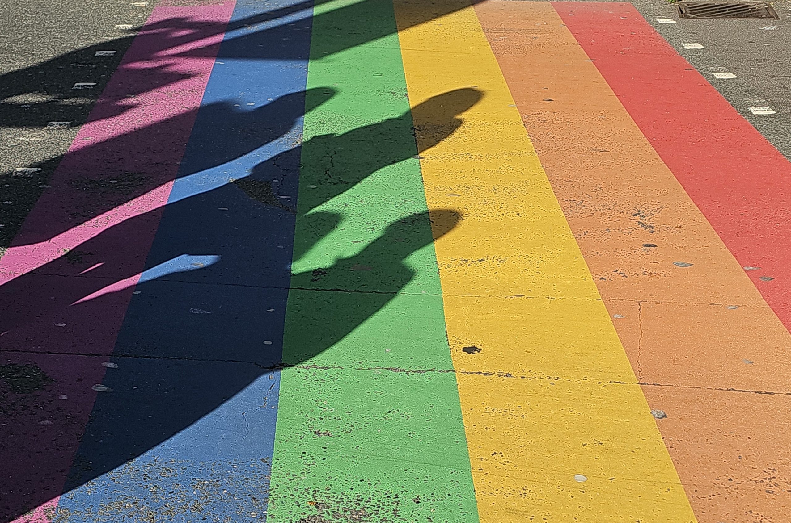 Pride flag painted on tarmac with shadows of passers by over it.