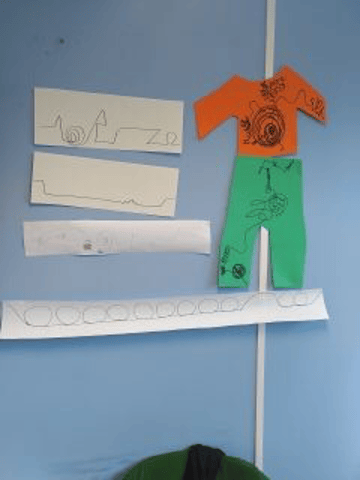 A top cut out of red card, trousers cut out of green card. Both are pinned to the wall. There are four other bits of cardboard stuck to the all with squiggles drawn on.