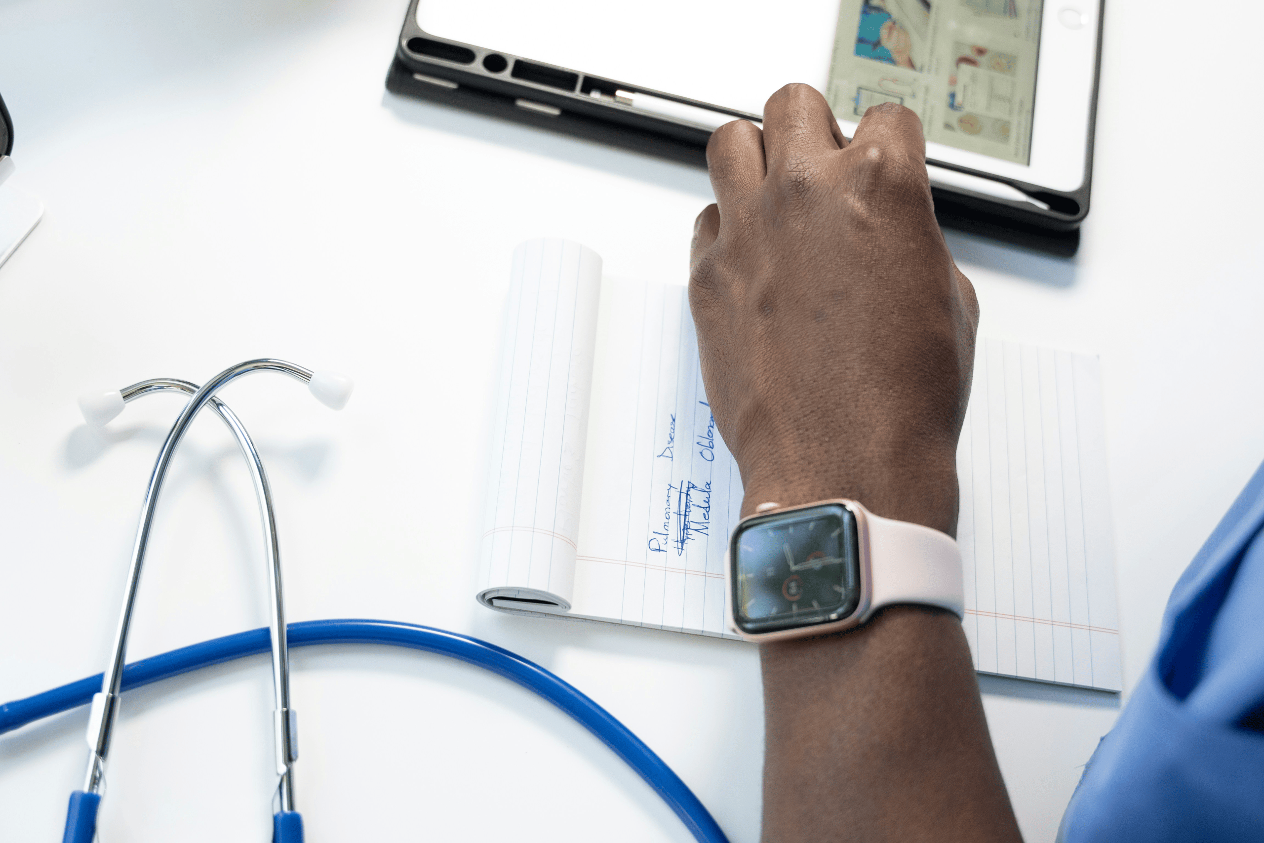The dark skin coloured hand of a medical professional resting on a notepad. There is a glimpse of a blue shirt which could be scrubs and a stethoscope laid out on a white table.