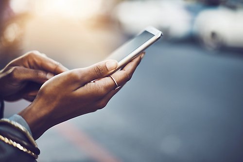 Image of a woman's hands holding a smartphone on city street, with a blurred line of parked cars in the background.