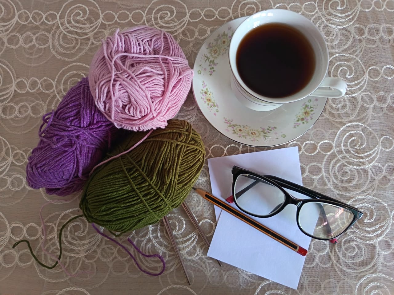 Photograph showing a cup of black tea/coffee, 3 balls of wool (pink, purple and dark green), some small squares of paper, a pencil and a pair of glasses. The items are arranged neatly on a table, covered by a lace tablecloth.