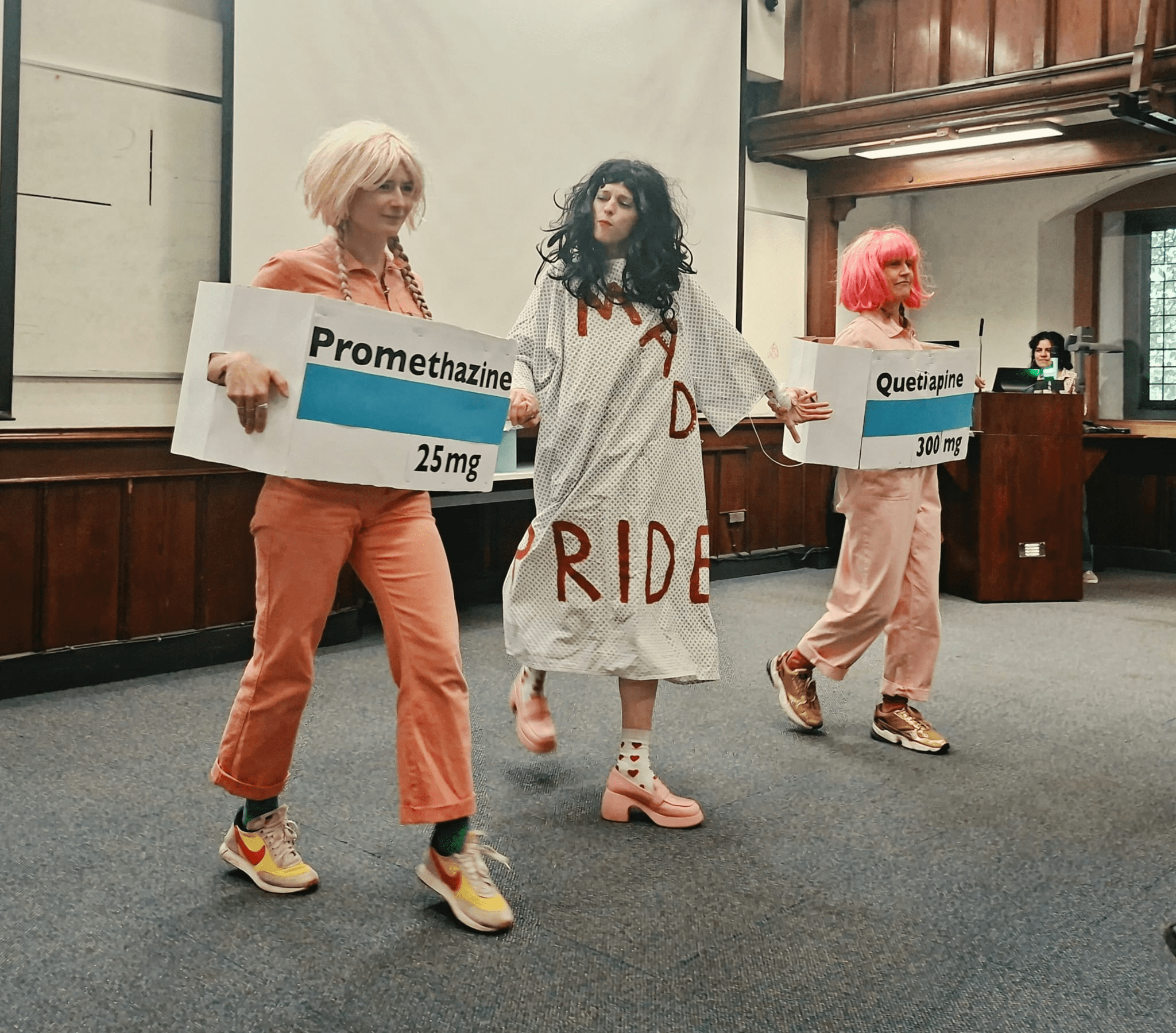 Photograph of three people in costume walking indoors. The person in the middle, wearing a dressing gown with ‘Mad Pride’ written on it, is flanked by two others holding large signs displaying medication names and dosages: “Promethazine 25 mg” and “Quetiapine 300 mg.”