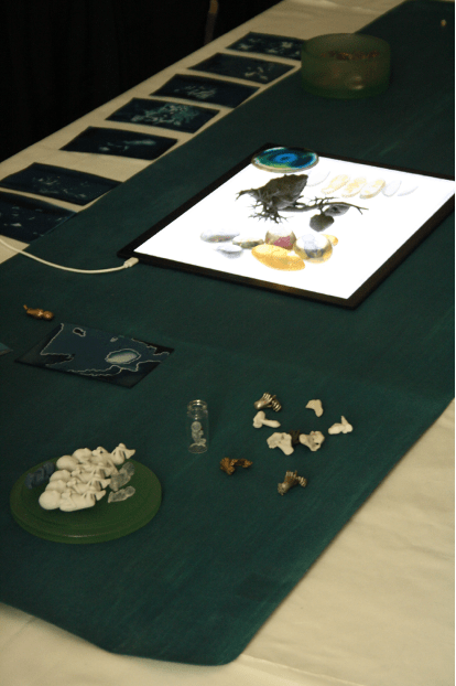 Close up photograph of objects on a dark green cloth on the table. What look like small plastic models of babies, small bones, and a lit ipad with a picture of eggs on it.
