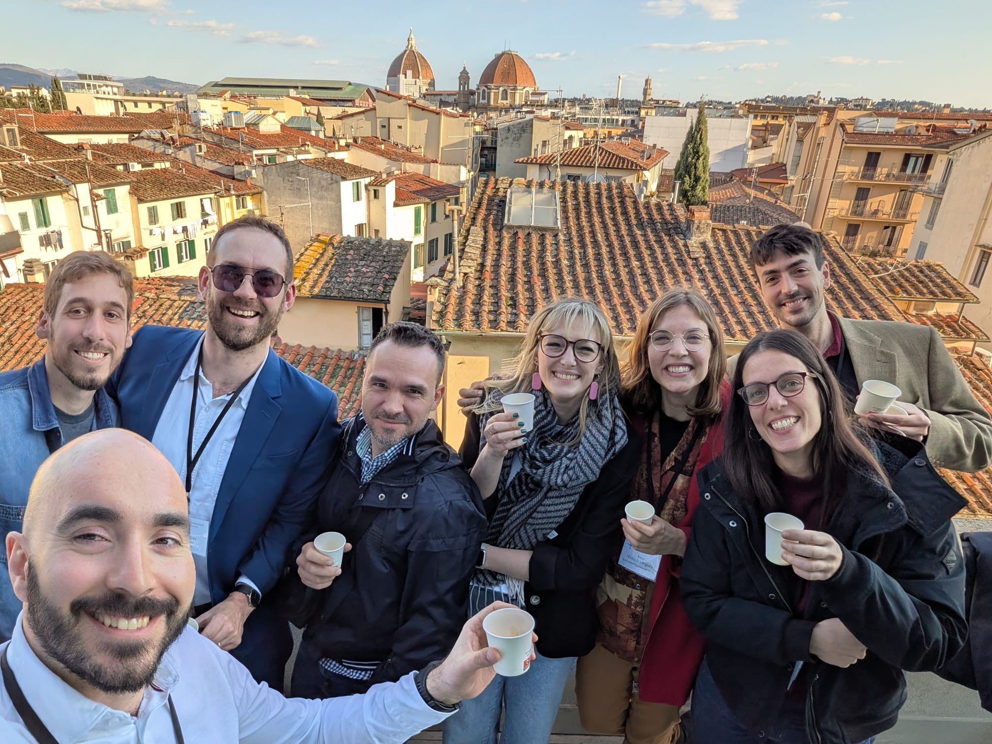 Group of people smiling and holding cups of coffee or water. Mediterranean city in the background.