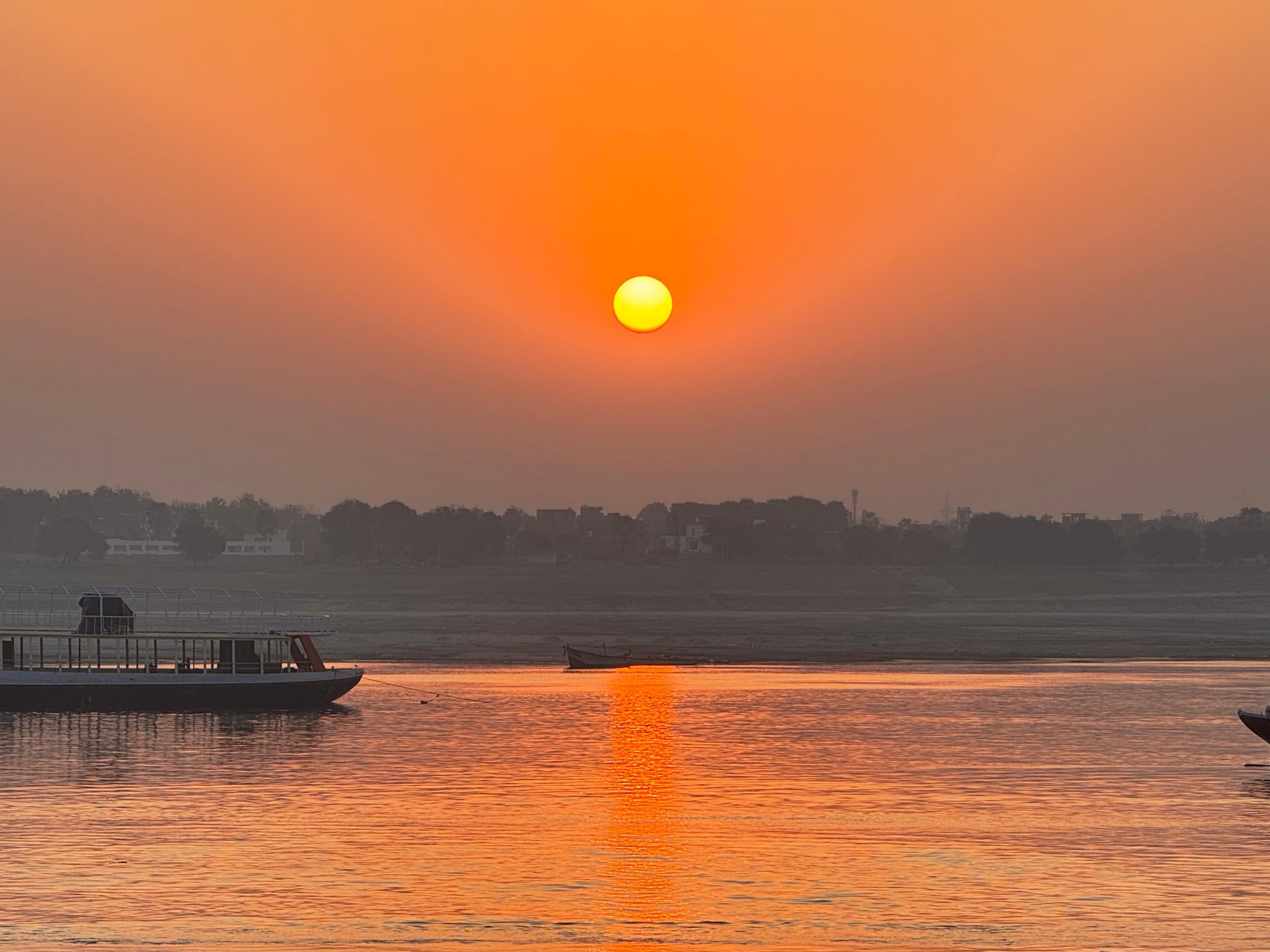 A photo of a sunset seen across a wide river in which long passenger boats move across the frame. The sun is centred and bright orange, with varying shades of orange radiating out across the landscape and reflecting in the calm water. On the other bank can be seen the vague and misty outline of a row of trees and the buildings of a city behind it.