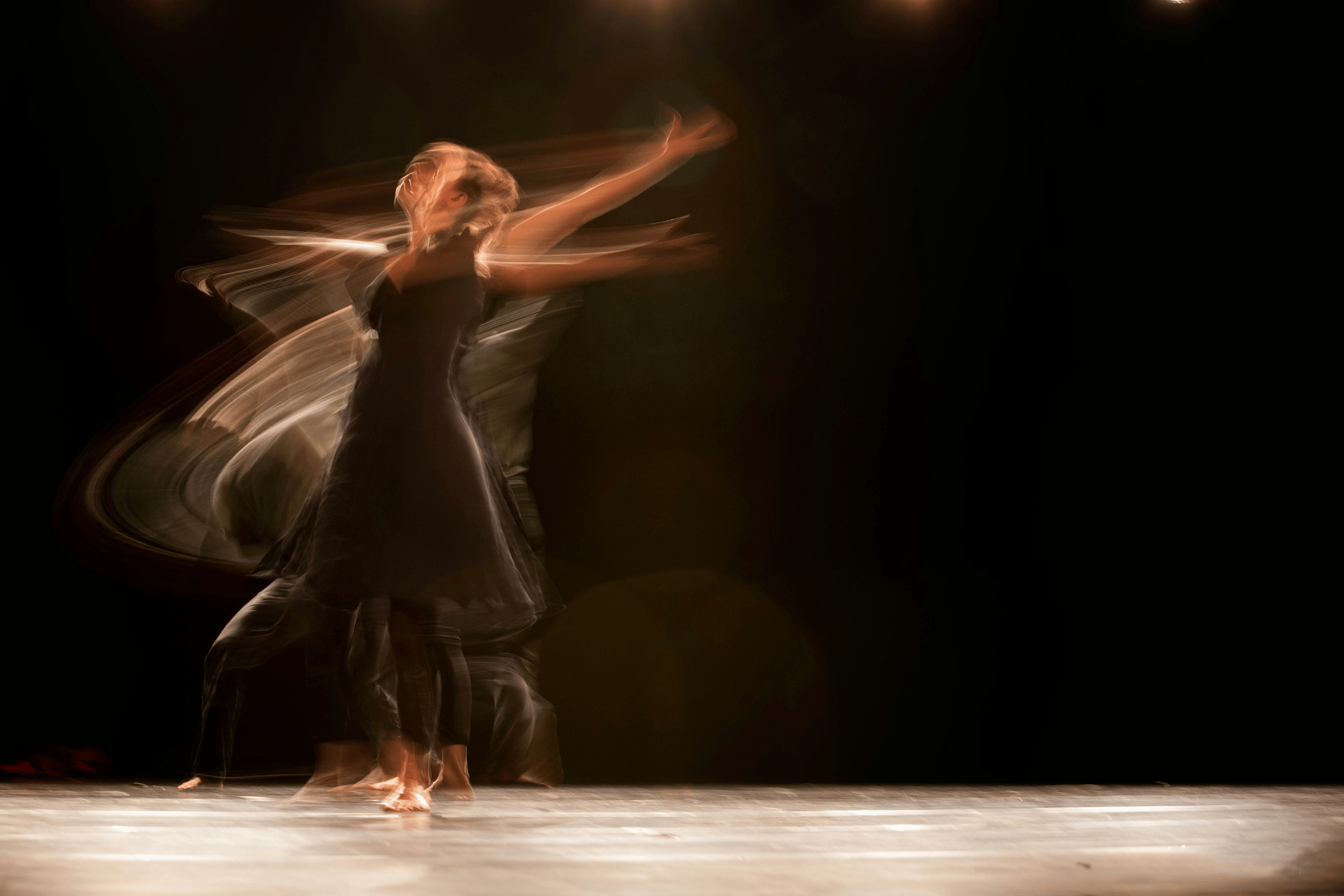 Image of a moving dancer the traces of blur indicating movement. The figure is illuminated against a black background.