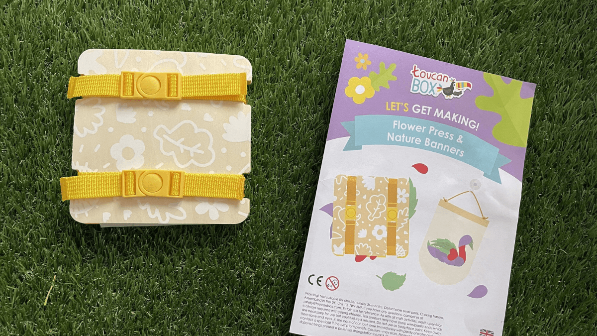 On a grass surface is a small wooden flower press with yellow straps. There are toucanBox instructions next to it