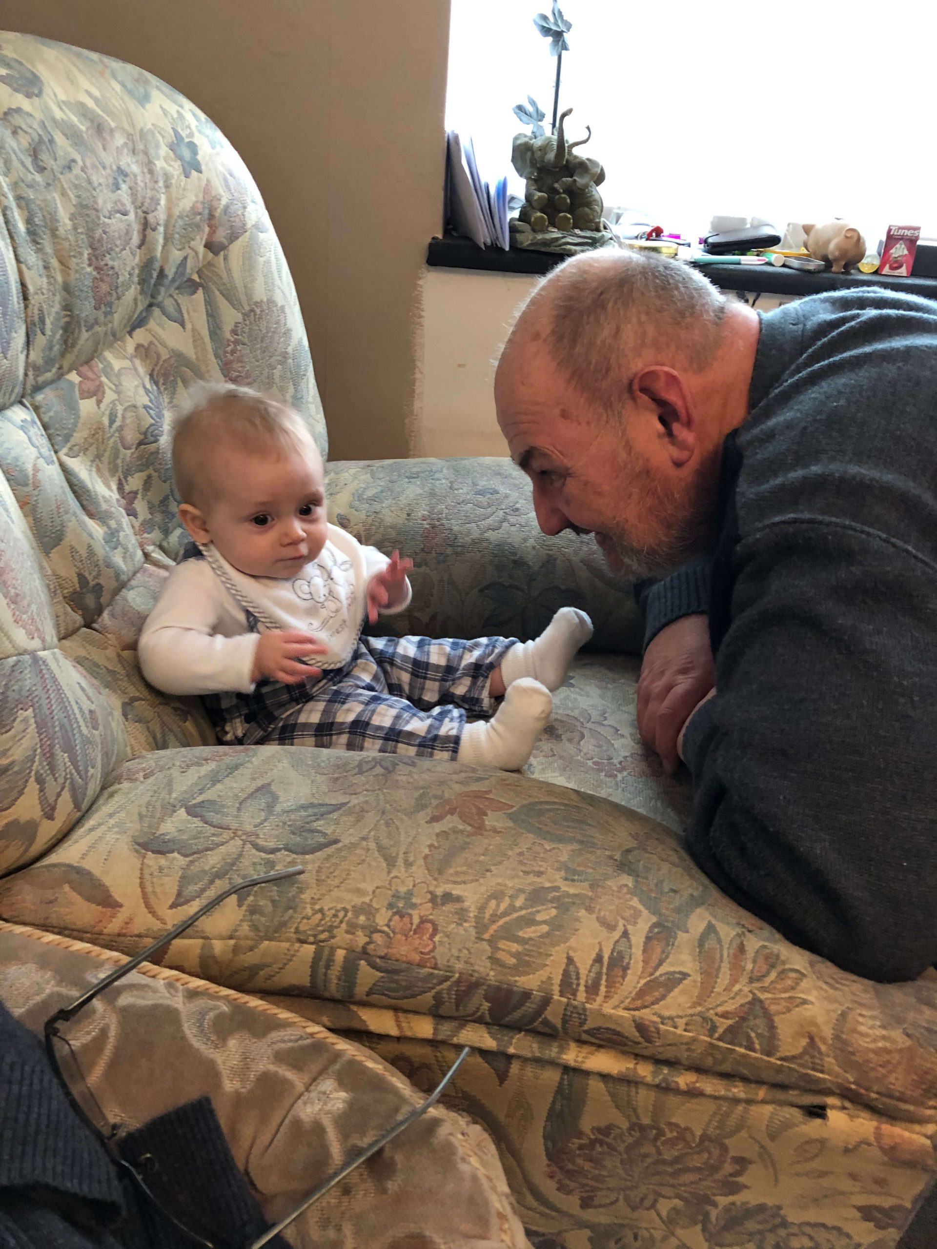 Baby J is sat on an armchair. He is wearing checkered dungarees and grandad is knelt on the floor in front of Baby J, playing with him