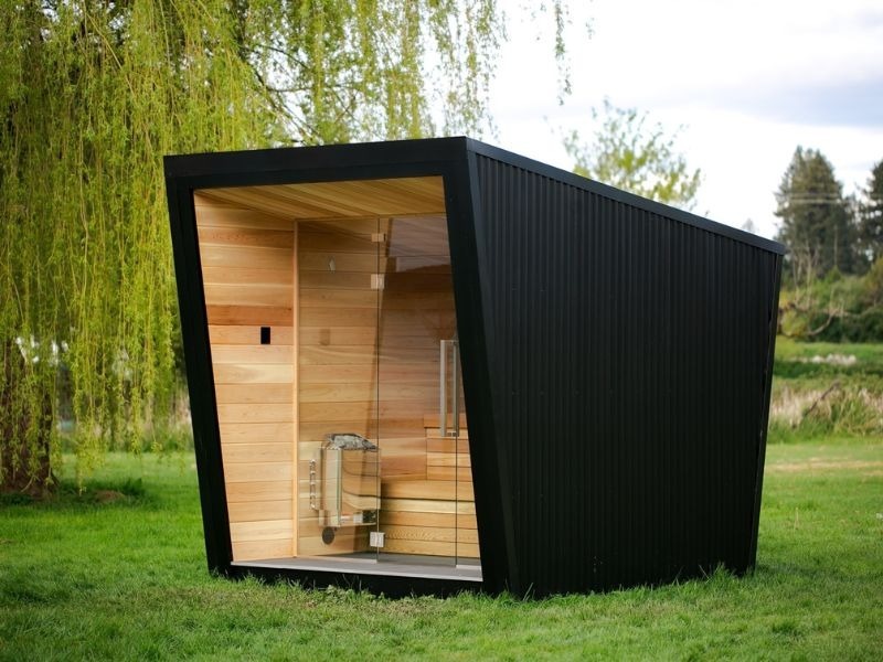Outdoor home sauna on a grassy site with a black exterior, glass door, warm wood interior, and visible electric sauna heater, showing site prep and electrical planning for installation.