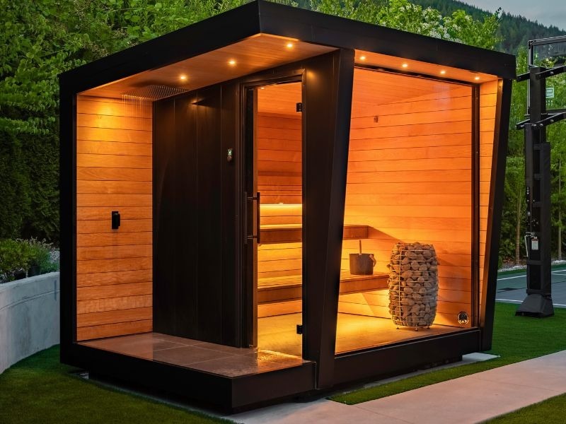 A modern black outdoor sauna with large glass panels, warm wood interiors, soft lighting, a visible sauna heater, and a sheltered entry area set within a landscaped backyard.