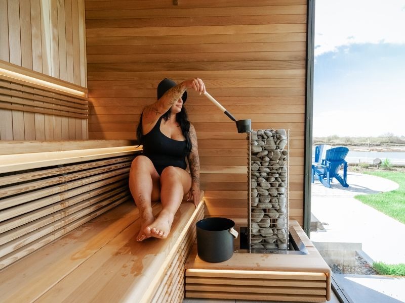 Woman pouring water over sauna stones inside a luxury wood sauna with tiered benches, warm lighting and an outdoor view.