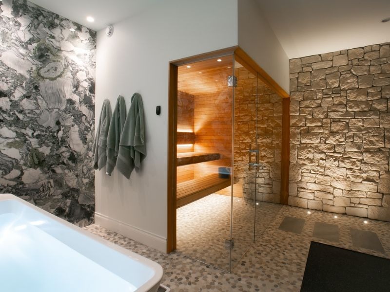 Built-in home sauna with glass enclosure integrated into a luxury bathroom with stone walls and a freestanding bathtub.