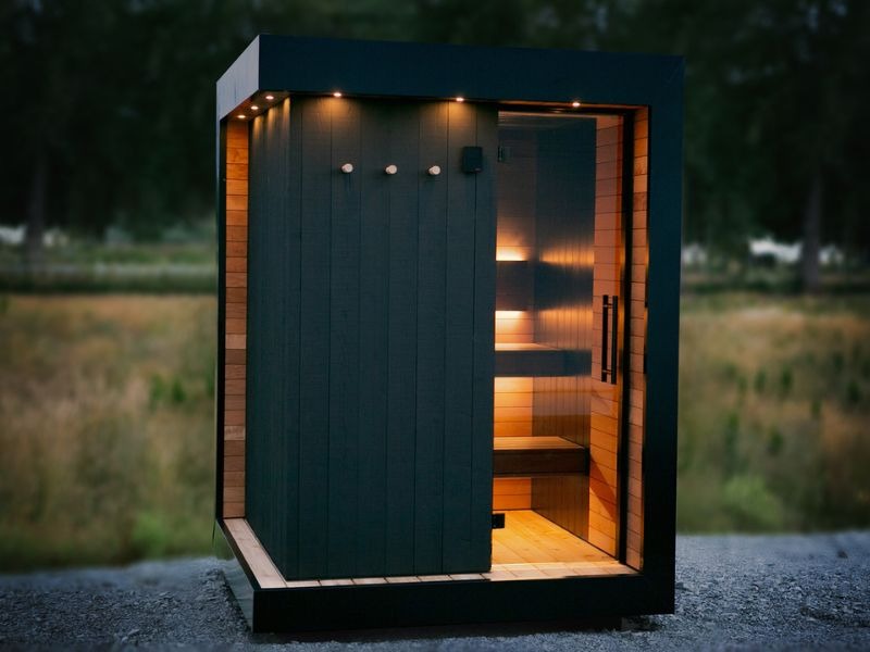 Modern black outdoor sauna illuminated at dusk with warm interior bench lighting, glass entry and integrated exterior downlights highlighting the sauna architecture.