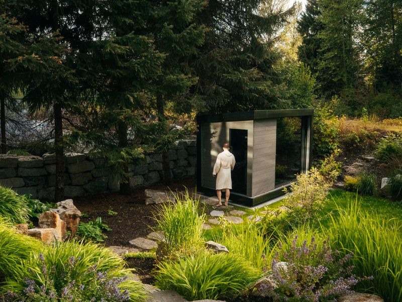 Person in a light robe entering a compact modern outdoor sauna with dark framing, wood cladding and full-height glass, surrounded by mature trees and landscaped greenery.