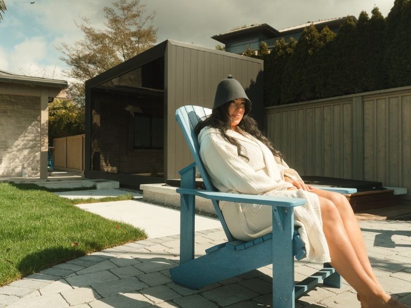 Luxury black outdoor sauna with full glass frontage in a Vancouver backyard with wet stone paving, covered lounge, fire feature, mature evergreen trees and lush privacy landscaping.