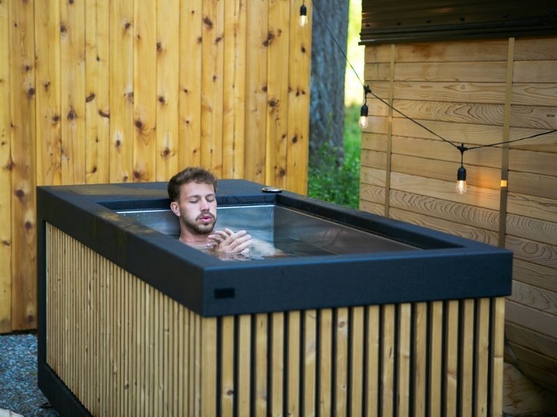 Man immersed to his shoulders in a rectangular outdoor cold plunge with a black rim and vertical wood slat exterior beside a wood-clad sauna and privacy fence.