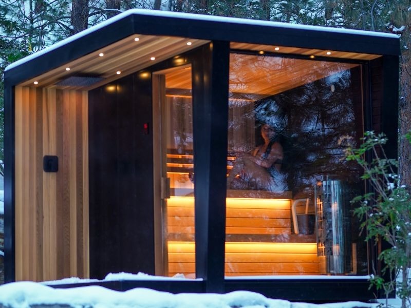 Modern black outdoor sauna glowing warmly in a snow-covered wooded setting, with full-height glass walls, illuminated wood benches and a person seated inside.