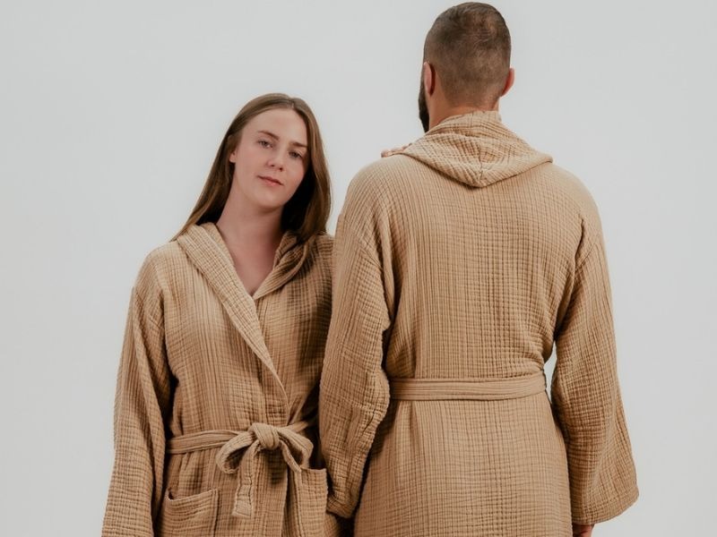 Two people modelling textured brown sauna robes with hoods, waist belts and front pockets against a light studio background.