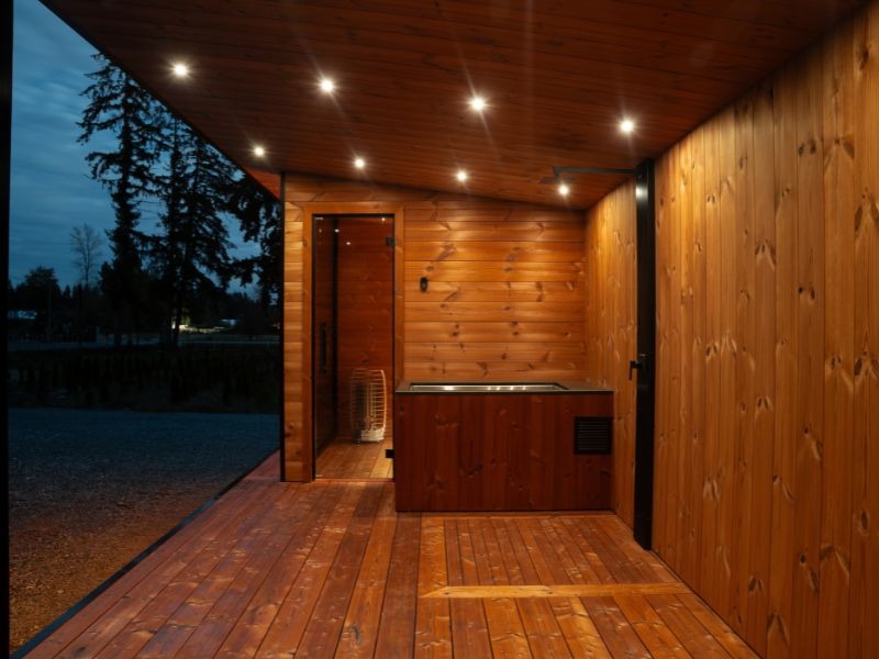 Covered outdoor sauna area with warm wood cladding, recessed ceiling lights, a glass sauna entrance and built-in wellness features at dusk