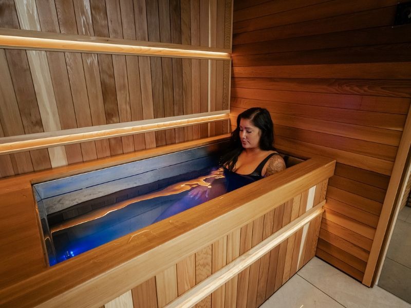 Woman immersed in an indoor cold plunge surrounded by warm wood panelling, integrated lighting and tiled flooring in a dedicated home wellness space.